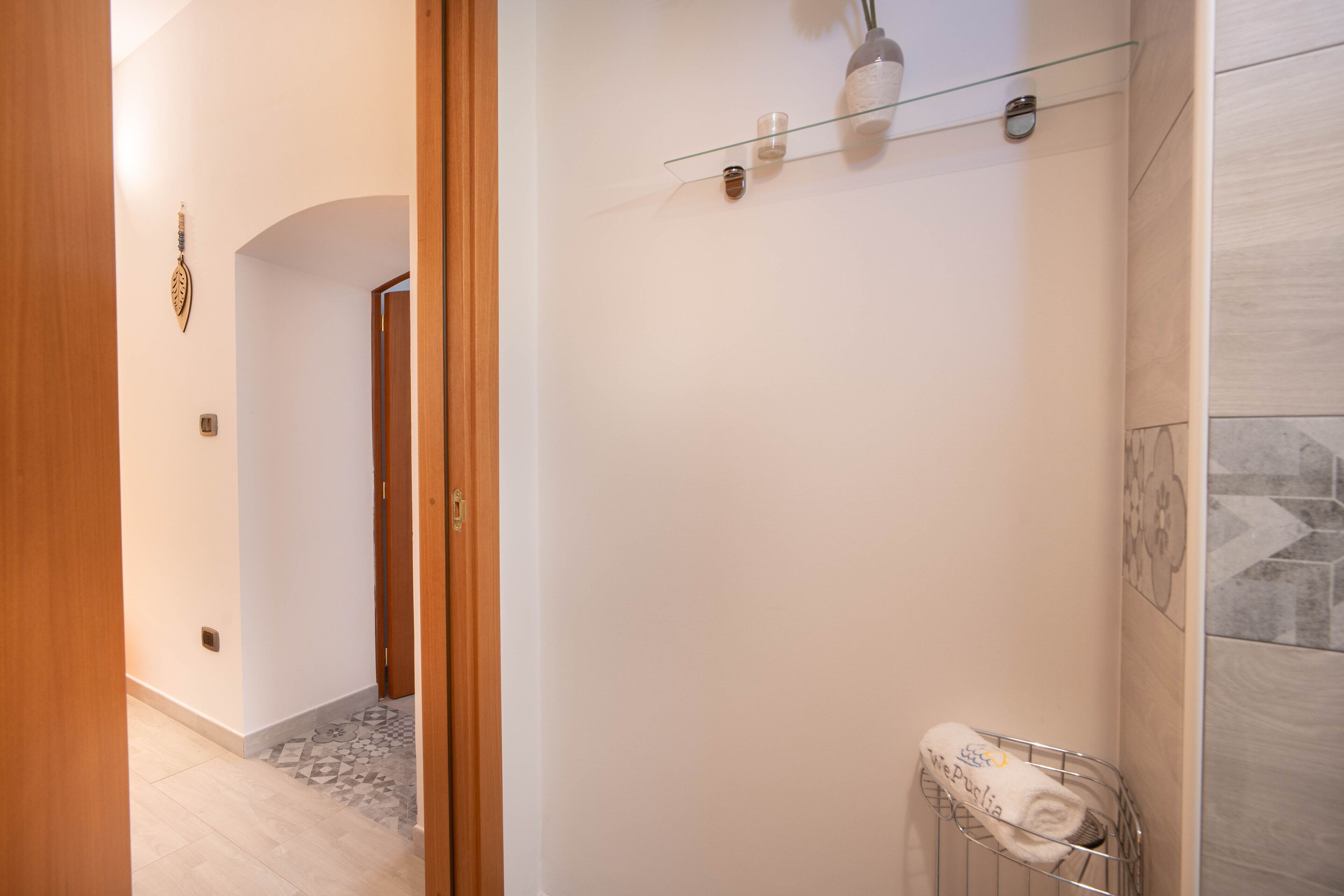 A bathroom with a glass shelf holding a towel and a glass vase, a tiled shower area, and a wooden door leading to another room.