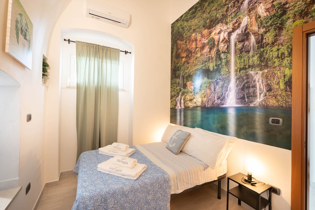 A bedroom with a bed, a nightstand, a wall-mounted air conditioner, a window with a curtain, and a large mural of a waterfall.