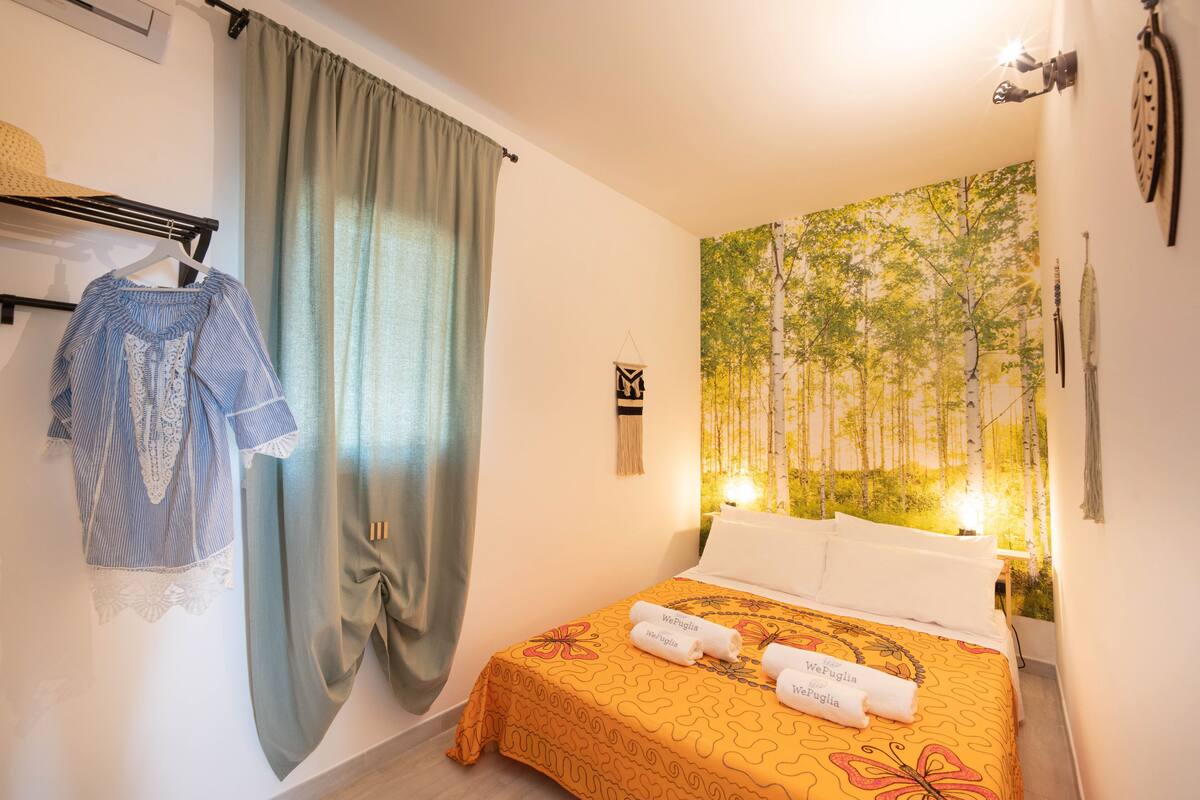 A hotel room with a bed, a wall tapestry, a hanging rack with clothes, and a curtain.