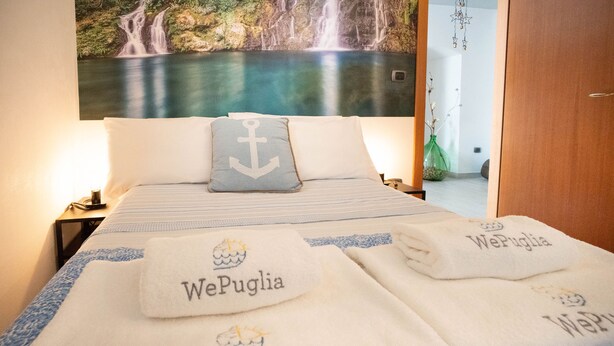 Habitación de hotel con cama y ropa de cama blanca y azul, almohada con diseño de ancla y toallas con el nombre 'WePuglia' bordado.