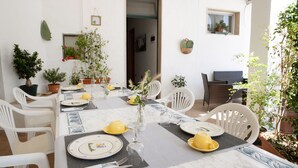Un comedor con una mesa dispuesta para cuatro personas, rodeada de sillas blancas, plantas en macetas y un ventanal.