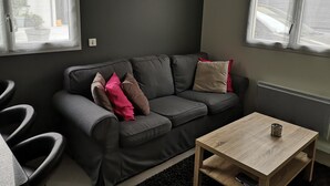 A modern living room with a grey sofa, a wooden coffee table, and a view of the street through large windows. - 1 Bedroom Apartment in Fresnes (Fresnes)