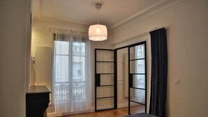 Une chambre à coucher comprenant un lit, une table de chevet, un luminaire suspendu, une fenêtre avec des rideaux légers et une porte-fenêtre coulissante donnant sur un balcon.