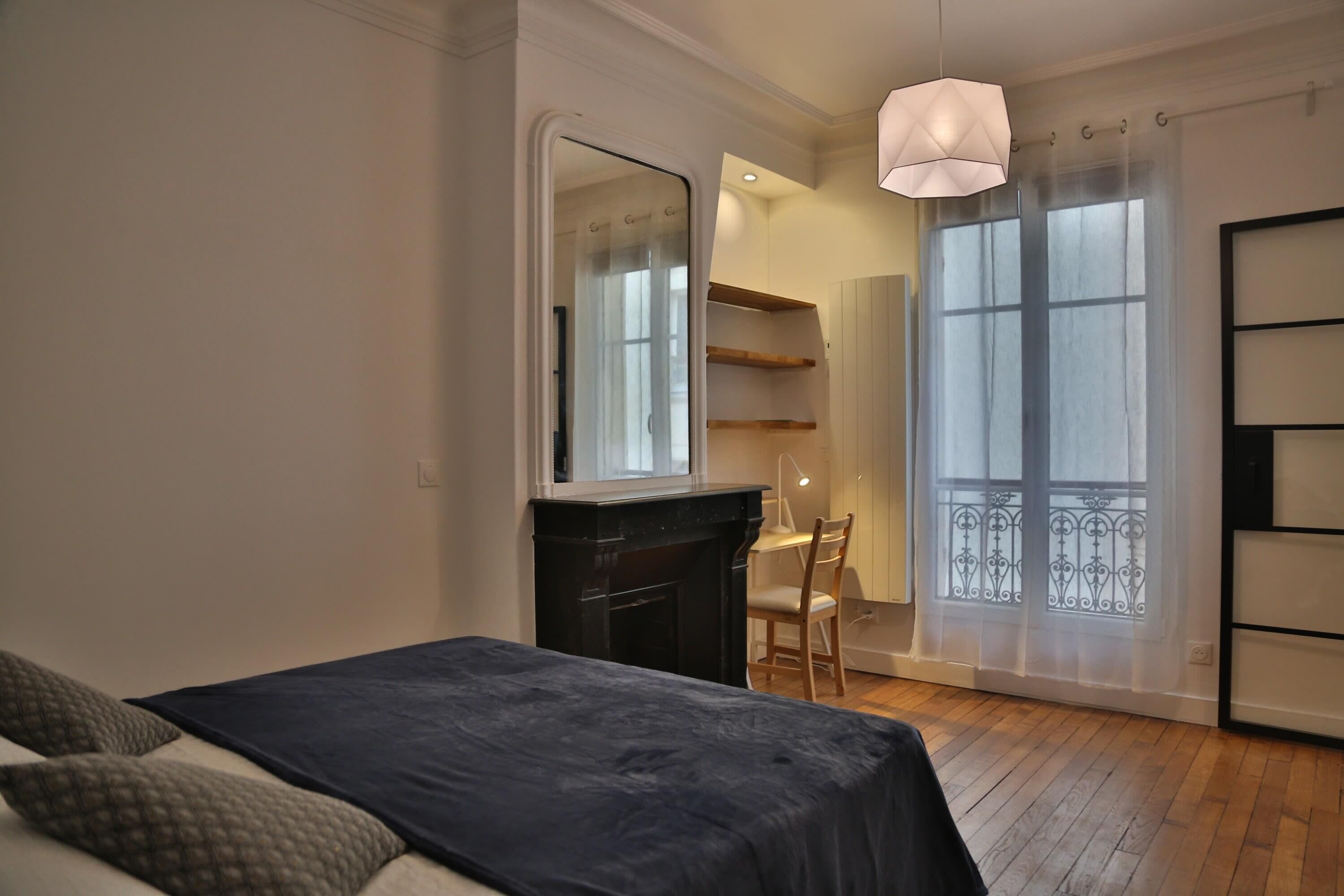 A bedroom with a bed, a desk, a chair, a fireplace, and a large window with a view.