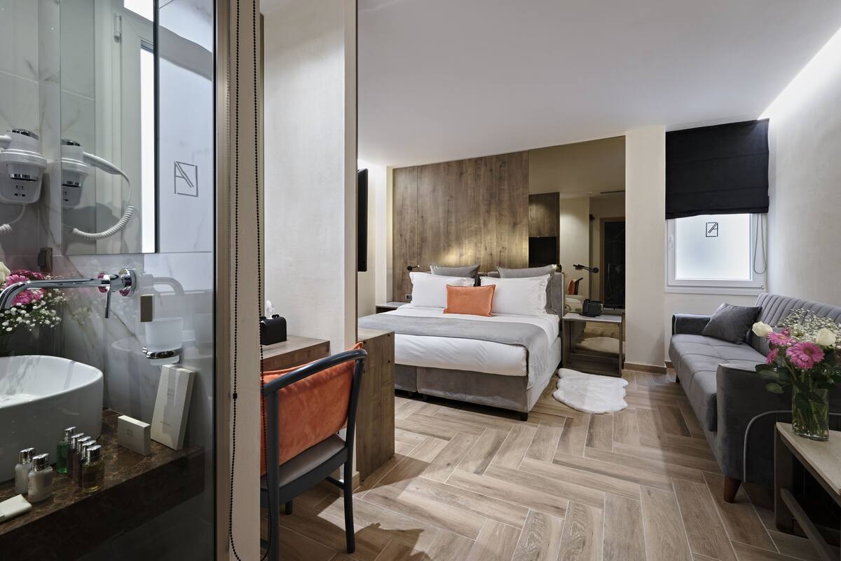 A modern hotel room with a large bathroom, a bed with white bedding, a grey sofa, and a wooden floor.