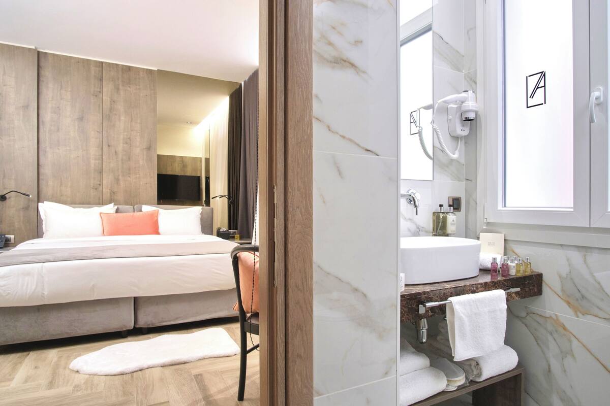 A modern hotel room with a large bed, a wooden wardrobe, a marble bathroom with a sink and mirror, and a robe rack.