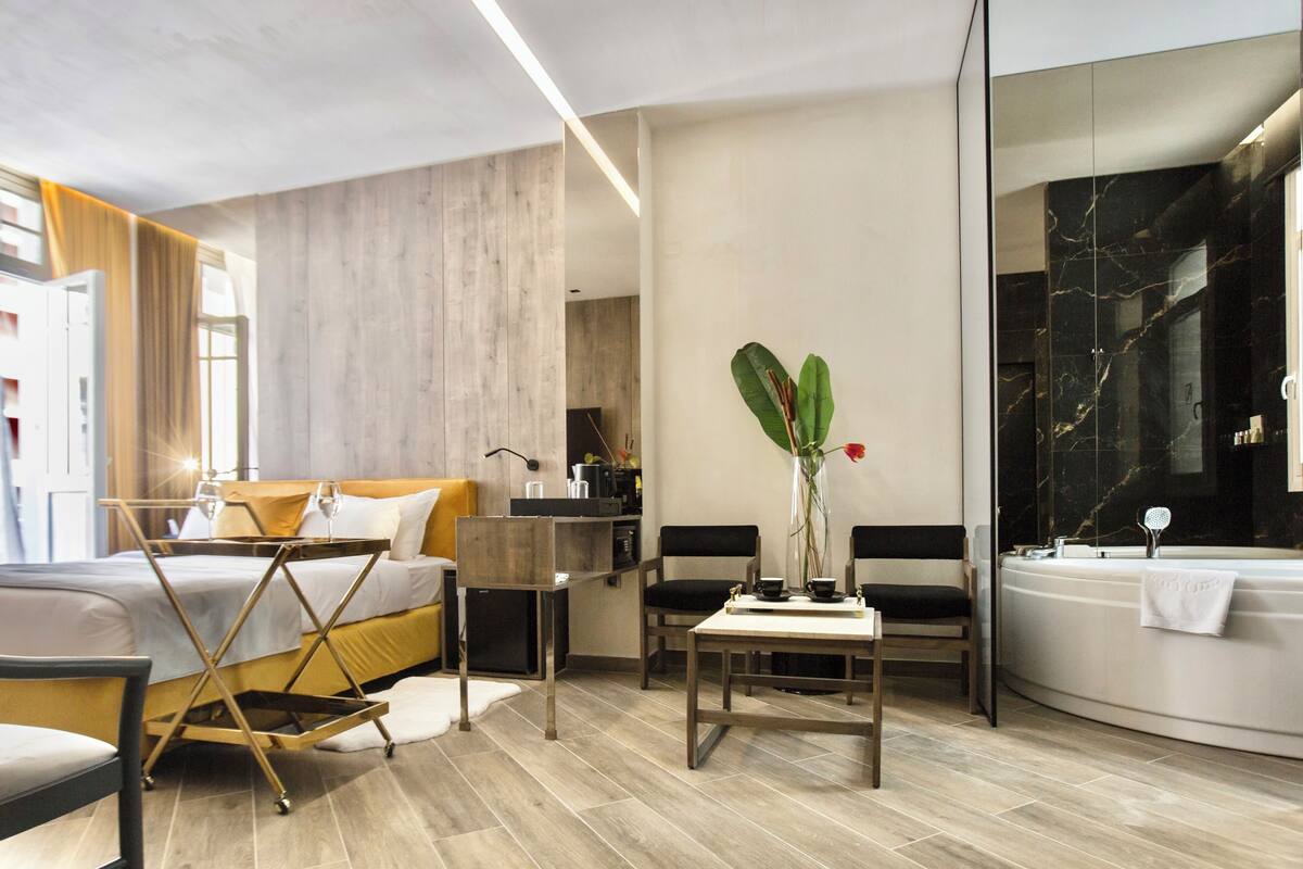 A modern hotel room with a bed, a small table, a chair, a desk, a bathroom with a bathtub, and a large mirror.