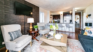 A modern living room with a sofa, armchairs, a coffee table, and a TV mounted on a dark wooden wall. - Cosmopolitan Condo - Lagoon Centre 701 In the Heart of Hilo (Hilo)
