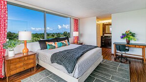 A bedroom with a large window, a bed with multiple pillows, a nightstand, a desk with a chair, and a potted plant. - Cosmopolitan Condo - Lagoon Centre 701 In the Heart of Hilo (Hilo)
