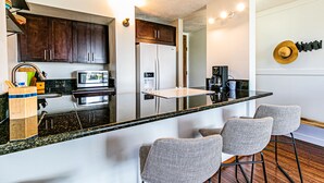 A modern kitchen with dark wooden cabinets, a stainless steel refrigerator, a microwave, and a coffee maker. There are three bar stools and a small island with a glass top. - Cosmopolitan Condo - Lagoon Centre 701 In the Heart of Hilo (Hilo)