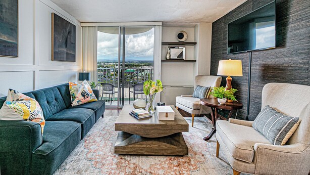 A modern living room with a sofa, armchair, coffee table, and a view of the city through large windows. - Cosmopolitan Condo - Lagoon Centre 701 In the Heart of Hilo (Hilo)