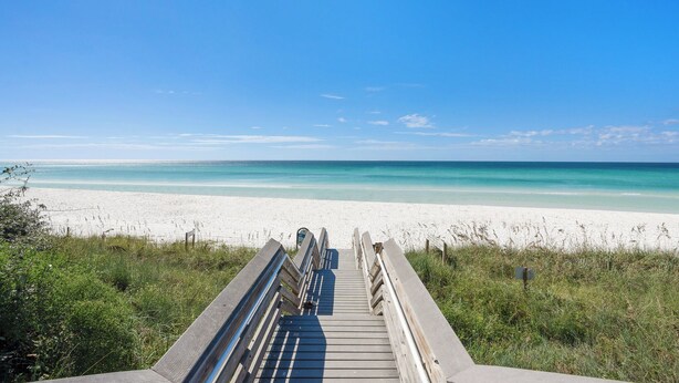 Een houten looppad dat naar een strand leidt met helderblauw water en wit zand.
