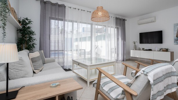 Un salon moderne avec un canapé, une table à manger et deux chaises. Un téléviseur est fixé au mur et une plante se trouve sur le côté gauche.
