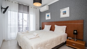 Une chambre d’hôtel avec un grand lit, deux tables de chevet, une petite table, une lampe et une fenêtre avec des rideaux légers.