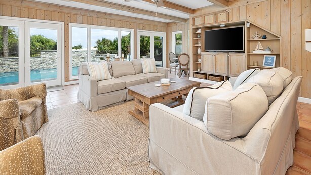 A spacious living room with a large sofa set, a coffee table, and a TV. The room has wooden walls and a ceiling fan. - Oceanfront w/ Private Pool & Rooftop Deck | Amazing Views | Sleeps 12 (Jekyll Island)