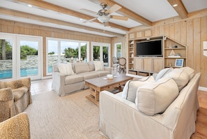 A spacious living room with a large sofa set, a coffee table, and a TV. The room has wooden walls and a ceiling fan.