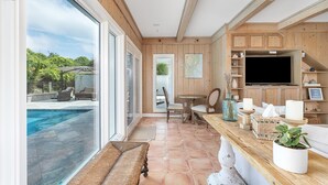 A spacious room with a large window overlooking a swimming pool, wooden paneling, a dining area with a table and chairs, and a kitchen area with cabinets and a television. - Oceanfront w/ Private Pool & Rooftop Deck | Amazing Views | Sleeps 12 (Jekyll Island)