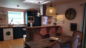 A modern kitchen with a central island, a dining area with a wooden table and wicker chairs, and a large wall clock.