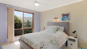 Une chambre avec une grande fenêtre, un lit recouvert d’une literie à motifs floraux, une table de chevet et un ventilateur de plafond.