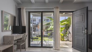 A bedroom with a bed, a desk, a chair, a TV, and a sliding glass door leading to an outdoor area with chairs and plants. - Adonis Luxury Village (Kassandra)
