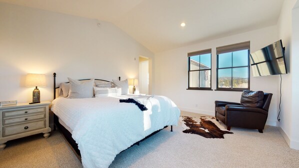 A bedroom with a large bed, a nightstand, a leather chair, and a TV mounted on the wall.