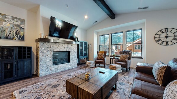 A spacious living room with a stone fireplace, large windows, a clock, and leather furniture. - Snowcreek VII 1403 Deluxe Townhouse, Stunning Mountain Views, 2 Car Garage, Washer Dryer by RedAwning (Mammoth Lakes)