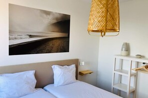 A hotel room with a bed, a wall-mounted shelf, a hanging lamp, and a large beach photograph. - In the Village - Madeira Surf Camp I (Machico)