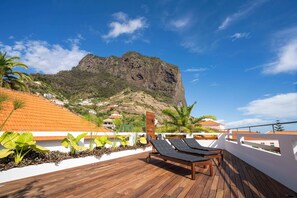 A wooden deck with lounge chairs, a white wall with plants, and a view of a mountain. - In the Village - Madeira Surf Camp I (Machico)
