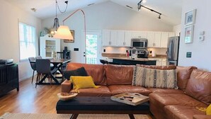 A modern living room with a brown leather sofa, a dining table with chairs, and a kitchen in the background. - CB House (Bike Trail, Brewery, Boardwalk, Park) (Carolina Beach)