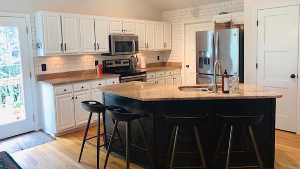 A modern kitchen with white cabinets, a large island with bar stools, stainless steel appliances, and a view to the outside. - CB House (Bike Trail, Brewery, Boardwalk, Park) (Carolina Beach)