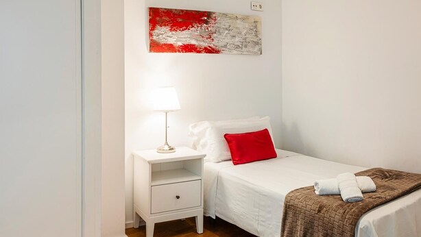 Un dormitorio ordenado con una cama individual, una mesita de noche con una lámpara y un almohadón rojo.