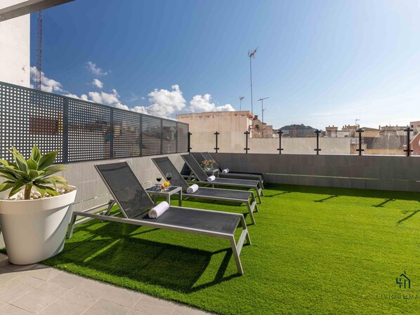 Eine Dachterrasse mit vier Liegestühlen, einem Topfpflanzen, einem kleinen Tisch mit einer Vase und Weingläsern.
