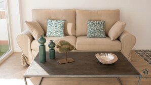 A beige sofa with patterned cushions, a wooden coffee table with decorative items, and a view of the outdoors through a window.