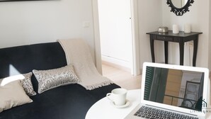 Una sala de estar moderna con un sofá negro, cojines estampados, un ordenador portátil sobre una mesa blanca, un cuadro enmarcado en la pared y un espejo negro ornamentado.