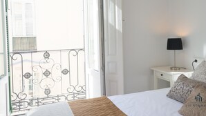 Habitación de hotel con una cama, una mesita de noche, una lámpara y un balcón con barandilla.
