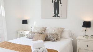 Una cama bien hecha con almohadones estampados, una toalla blanca enrollada y un plaid marrón. Hay dos mesitas de noche con lámparas y objetos decorativos.