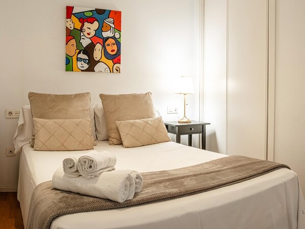 A neatly made bed with beige pillows and a rolled white towel, a small bedside table with a lamp, and a colorful abstract painting on the wall.