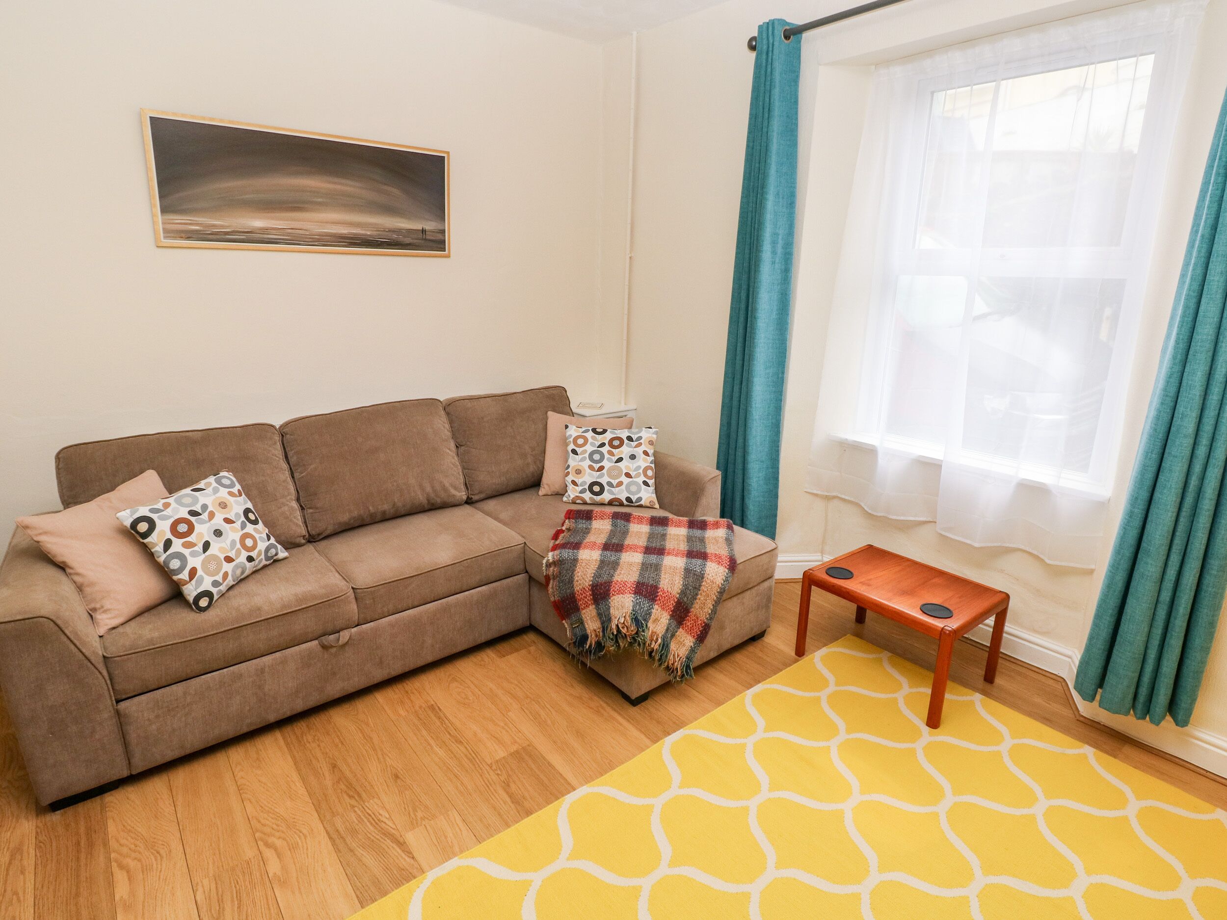 A living room with a brown sofa, patterned cushions, a plaid throw blanket, a yellow rug, a small wooden table, and a painting on the wall.