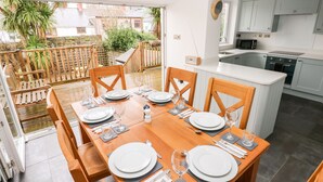 A dining area with a wooden table set for six, a kitchen with white cabinets, and a view of the garden through the glass door. - Ty Cwtch (Tenby)