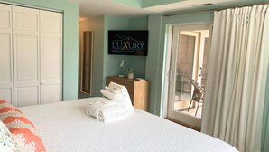 A hotel room with a large bed, a desk with a plant, a sliding glass door to an outdoor area, and a sign for 'LUXURY RENTALS OF HILTON HEAD'. - 237 Shorewood Villas - Ocean Views - Exceptional Update - Discounted Rates! (Hilton Head Island)
