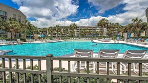 A large outdoor swimming pool with lounge chairs and palm trees, surrounded by a multi-story building.