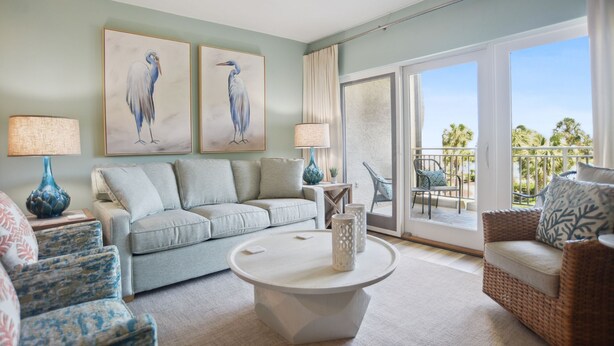 A living room with a light grey sofa, a round coffee table, and two blue vases. There are two paintings of birds on the wall, and a balcony with outdoor furniture. - 237 Shorewood Villas - Ocean Views - Exceptional Update - Discounted Rates! (Hilton Head Island)