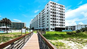A modern hotel with multiple floors, a wooden boardwalk leading to the entrance, and a clear blue sky. - Flip Flop O'Clock-Gulf Views • 6 Pools • Beachfront Resort Escape (Gulf Shores)