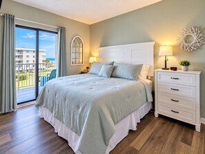 A bedroom with a large bed, a nightstand with a lamp, a wall clock, and a view of a building through a window.