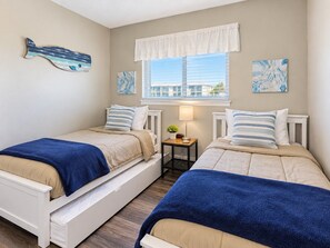 A bedroom with two beds, a window with blinds, a bedside table with a lamp, and artwork on the walls.