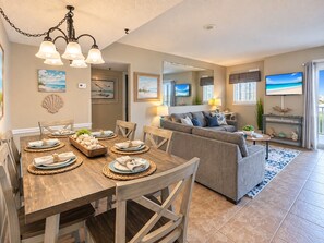 A modern living room with a gray sofa, a dining area with a wooden table set for four, and a large flat-screen TV mounted on the wall. - Gulf View Getaway | Beach + Heated Pool | Sleeps 6 (Gulf Shores)