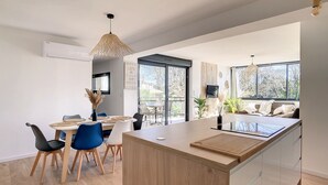 Una cocina moderna con una isla central, zona de comedor con sillas y una mesa, y ventanas amplias que dejan entrar luz natural.