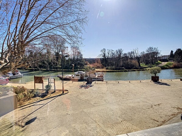 A riverside park with a concrete walkway, benches, and a boat dock. - View of the Canal du Midi - Comfortable apartment for 6 people (La Redorte)