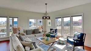 Ein geräumiges Wohnzimmer mit einer Couch, Sesseln, einem Couchtisch und einer Deckenleuchte. Große Fenster bieten einen Ausblick nach draußen.