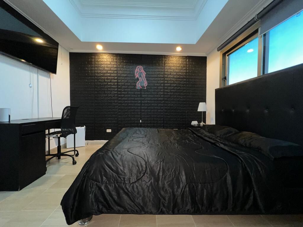 Un dormitorio con cabecera negra, cama con cubierta negra, escritorio con silla y un letrero de neón en la pared.